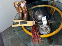 Wide view of all lot items on glass table: cowboy boots, leather helmet with ear flaps, boot jack, metal bowl, and horsetail fly whisk