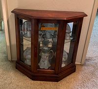 Full view of vintage cherry wood curio cabinet with glass front and angled glass sides. Displays porcelain items inside. Measures 30.5" wide x 11.25" deep x 29" high shown on orange tape inside.