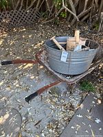 Full view of rusty metal wheelbarrow holding galvanized steel tub containing wooden watering cans.