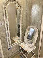 Tall white wicker framed rectangular mirror set on floor leaning against wall with smaller oval mirror and stool in the foreground.