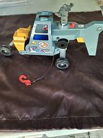 Side view of gray Ghostbusters helicopter toy showing logos, yellow rear engine section, black wheels, and red hook attached to string.