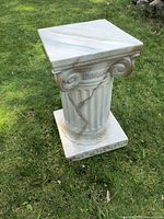 Ceramic Roman-style column pedestal shown standing on grass outdoors, showing fluted shaft, volutes on the capital, patterned square base and marbled ceramic finish.