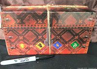 Rectangular decorative box with vintage design and Hogwarts house symbols, secured with string and clasp, with a Sharpie marker for size reference.