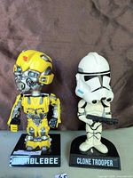 Two bobble-head figures. Left is Bumblebee from Transformers with yellow and gray details and blue eyes. Right is a Star Wars Clone Trooper figure in white armor holding a blaster. Both are mounted on bases labeled with their character names.