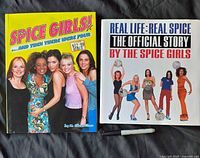 Two hardcover Spice Girls books arranged side by side on a black cloth surface: one titled 'Spice Girls! ...and then there were four' with a photo of the group on a yellow cover and a price sticker visible, the other titled 'Real Life: Real Spice The Official Story By The Spice Girls' with a white cover featuring the five members in colorful outfits.