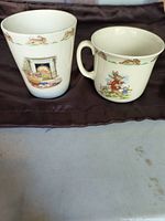 Two cream-colored Royal Doulton Bunnykins ceramic pieces placed side by side on a brown fabric with a plain surface underneath, showing the bunny and fireplace illustrations clearly.