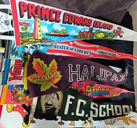View of seven vintage fabric pennants in varying theme and colours, showing printed graphics and text including Prince Edward Island lighthouse, Statue of Liberty, Canada maple leaf, Halifax parade, and F.C. School text.