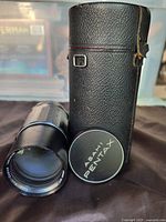 Photo showing Asahi Pentax black camera lens, lens cap with Asahi Pentax logo, and black textured leather carrying case with buckle.