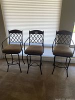 Three wide seat bar stools with metal frames and cushioned beige seats, positioned side by side in front of window blinds.