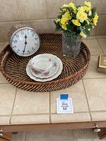 Woven tray holding round wall clock, ceramic tea set, and vase with yellow artificial flowers on tiled countertop