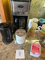 Full view of all items on kitchen counter including Cuisinart coffee maker, black kettle, white coffee grinder, Aerolatte frothier, multiple mugs, and sugar packets.
