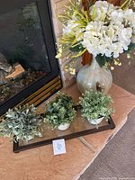 Photo showing one large faux floral vase arrangement with white flowers and three smaller faux potted plants on a tray in front of a fireplace.