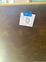 Close-up of worn brown surface of the folding card table with a Maxsold label attached.