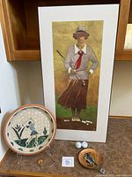 Golf related items arranged on a table including framed golf art print, mosaic golf plate, ceramic bird plate, two golf balls, and golf medallion