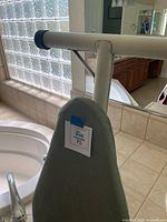 Side view of the ironing board standing next to a bathtub, showing part of the greyish blue cover and frame.
