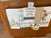 Four kitchen towels hanging on a wooden rack showing designs including rabbits, plaid pattern, humorous text, and Napa Valley branding.