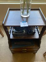 Front view of the wooden night stand showing top surface with vase, middle shelf holding a black clock radio and handheld barcode scanner, and lower drawers containing folders.
