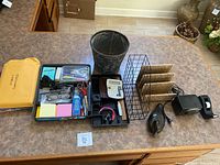 Overview of all office desk items grouped on brown counter consisting of organizer trays, tape dispenser, calculator, file sorter, sunglasses, and waste bin.