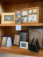 Multiple picture frames on shelf including wooden and black frames, with one painted vase and black book holders