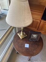 Round wood end table with brass lamp and decorative clock on top near window, showing overall composition and size