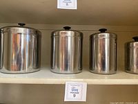 Shelf with four larger stainless steel canisters of varying sizes with black knob lids arranged from largest to smaller.
