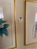 Two floral picture frames hung side by side on a beige wall, showing part of the artworks depicting blue leaves and white flowers in clear vases, gold frames visible.