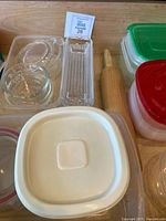 Various food storage containers with lids in red and green, glass bowls and rectangular glass dishes, wooden rolling pin on a table