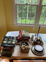 Overview of assorted table linens stacked, plates nested, decorative baskets with faux flowers, shaker, and kitchen timer arranged on table.