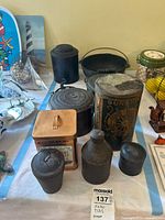 Full view of 7 assorted vintage metal tins and containers arranged on a blue and white cloth, including a large Sunrise Pilot Wafers tin and cast iron scotch bowl.