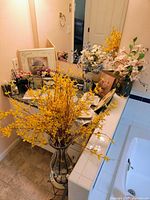 Photo showing a master bathroom counter and corner with an assortment of floral and ceramic decorative items including yellow faux flowers in a tall clear vase and various faux floral arrangements.