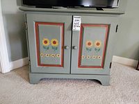 Front left angled view of green wood cabinet with sunflower designs on doors, showing overall piece and painted decorative details.