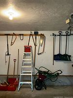 Wide shot of garage wall showing ladder, saws, gas cans, broadcast spreader, wheelbarrow, and other tools arranged on floor and hung on wall