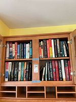 Books arranged inside a wooden cabinet with two shelves visible, featuring a mix of hardcover and paperback books in a range of themes.
