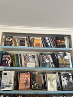 Three shelving units with approximately 60-80 books total in mixed bindings, various genres visible, arranged mostly upright with some stacked horizontally.