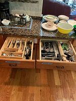 Wide view showing two kitchen drawers open with assorted cutlery and utensils neatly organized in wooden and metal trays, accompanied by various plates, bowls, cups on the countertop.