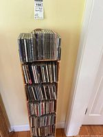 Full view of the wicker CD rack fully loaded with music CDs stacked across three shelves against a beige wall.