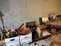 Wide view showing multiple cardboard boxes containing assorted garage supplies including automotive fluids, cords, and a socket set case.