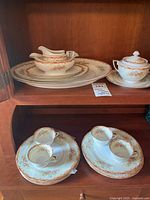 Top shelf of wooden cabinet showcasing gravy boat, covered sugar bowl, and multiple plates with floral pattern and gold trim.