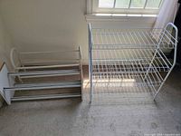 Two white shoe racks placed near a window on carpeted floor showing both metal and wood design shoe rack and one white wire tiered shoe rack.