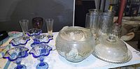 Photo showing vintage-style glass punch bowl with floral design, matching ladle, six blue-rimmed punch cups, assorted clear and tinted glasses, and glass cake stand with dome lid.