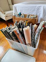 Numerous rolled posters stored upright in a translucent plastic bin and adjacent cardboard box on hardwood floor