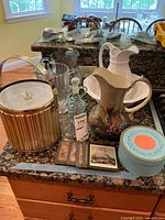 Photo showing assortment of pitchers including ceramic and glass, a gold-tone metal ice bucket with lid, a set of cocktail plates in original round box, and decorative coasters arranged on a kitchen counter.