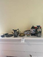 Wide view showing all items on shelf including metal teapot, cloisonné vase, ginger jar on wooden stand, small bowls and plates