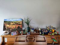 Wide view of a collection of assorted vintage decorative items arranged on a wooden piano top, including tile art, ceramic vases, terracotta figurines, and colorful animal figurines.