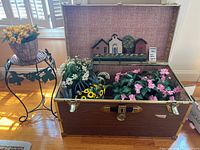 Photo of vintage style trunk open showing various artificial flowers, wall hangings inside lid, and metal plant stand next to it with basket of yellow tulips.