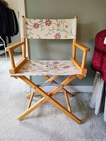 Front view of wooden director's chair with cream canvas seat and back printed with red, pink and green floral pattern, wooden arms and folding legs.
