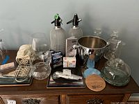 Full view of barware items arranged on table including siphon bottles, ice bucket, decanters, trivets, cheese plate, pitcher, accessories.