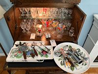 Wide shot of open vintage wooden cabinet with multiple styles of glassware (martini, brandy, cordial, shot glasses) on shelves. Two plastic serving trays on pull-out shelf with various metal bar utensils with wooden handles arranged on top.
