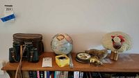 Front view of all items together on a wooden shelf including Kenko binoculars, leather case, French globe, various animal figurines, and round coasters