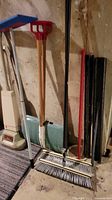 Photo showing several push brooms with wooden and metal handles, a red snow shovel, green snow shovel with metal edge, and black metal scraper leaning against a wall.